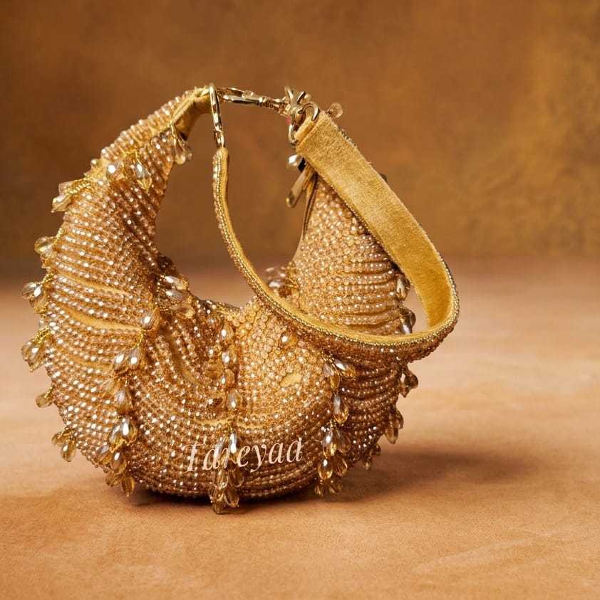 A gold-toned crescent-shaped beaded clutch bag with a wide matching strap, displayed on a textured brown surface against a matching brown background.
